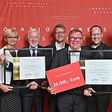 Personen stehen nebeneinander und halten eine Urkunde und einen Scheck